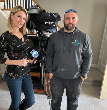 HVAC technician standing beside a news reporter with camera equipment inside a home