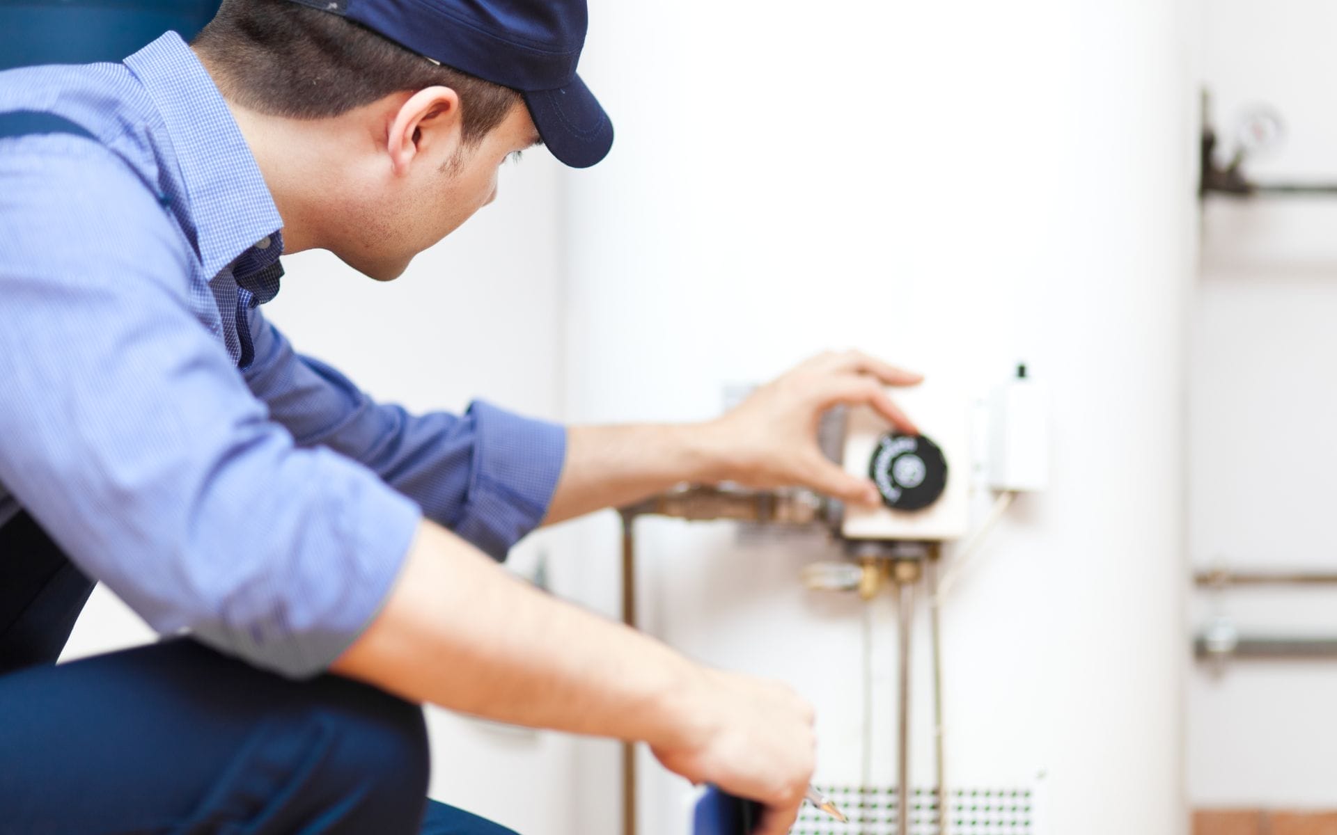 Technician kneels to service a wall-mounted water heater/boiler, adjusting plumbing connections with tools during a home repair visit.
