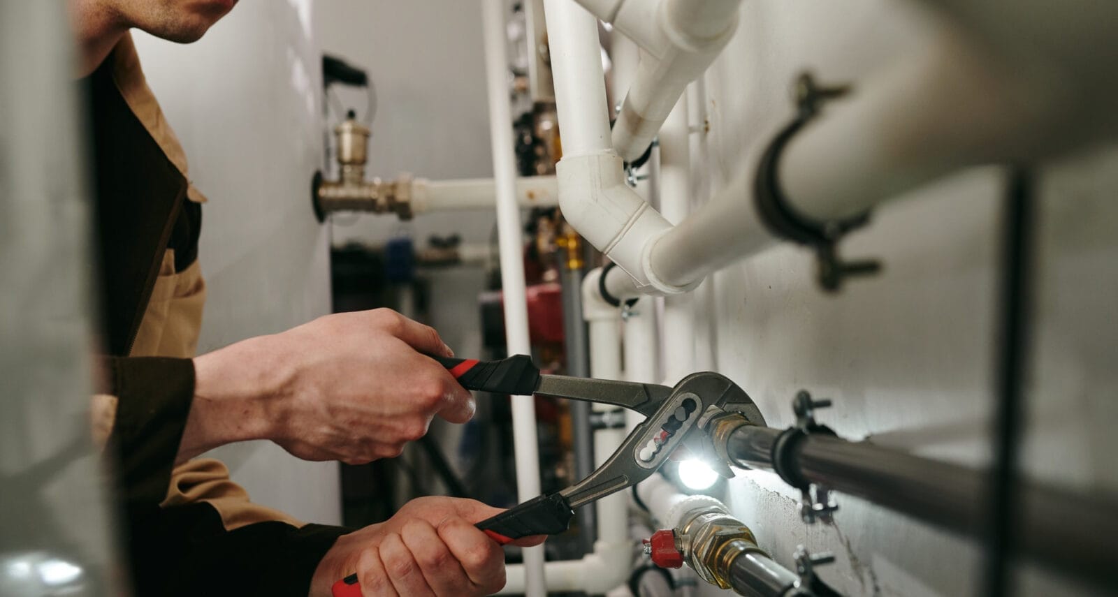 A plumber wearing a work uniform and safety gloves is crouched near exposed pipes, using a wrench to tighten a connection.