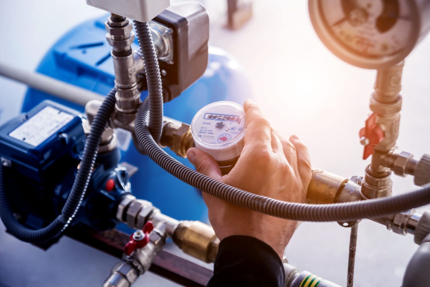 A close-up of a man's hand turning the dial of a water meter on a residential water system. The meter displays measurement readings, with surrounding pipes and valves visible in the background. The setting appears clean and well-maintained, with natural light highlighting the components.